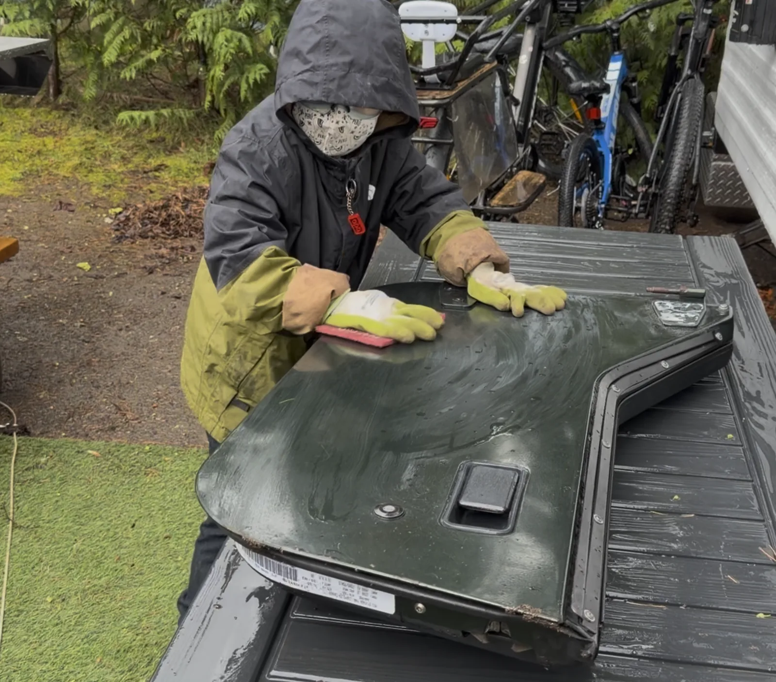 Craig in a mask and hoodie sanding the Jeep hood by hand on the grass