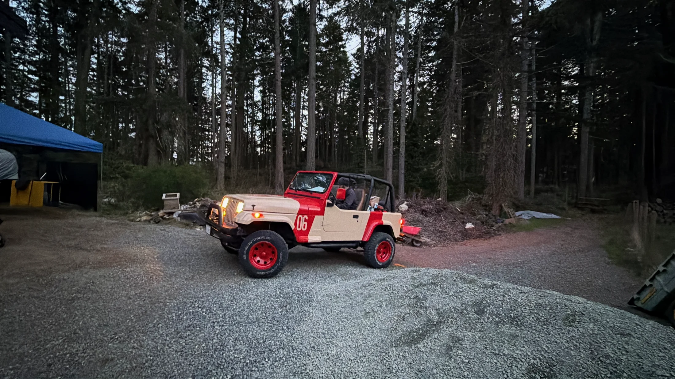 The finished Jurassic Park Jeep — a tan and red 1995 Wrangler YJ with a red '06' and red wheels, headlights on in a forest clearing